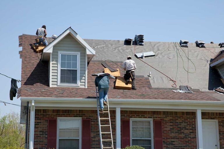 A person on a ladder cleaning leaves and debris out of a home’s roof gutter with gloves.