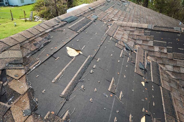 A fallen tree branch rests across damaged shingles beside bent gutters after a storm.
